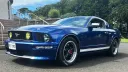 Blue Ford Mustang wedding car parked on a gravel driveway with chrome wheels and bonnet ribbons.