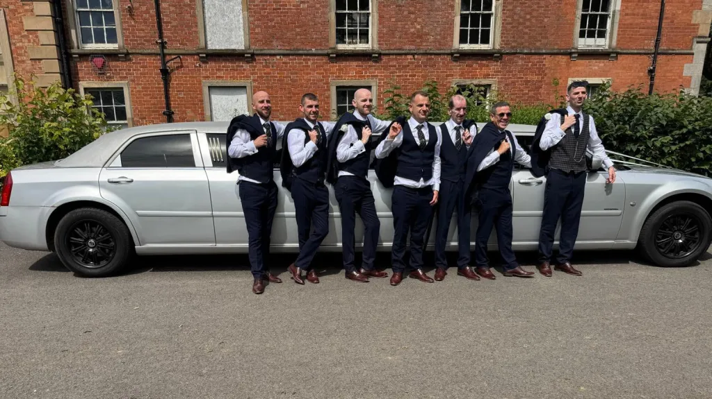 Group of groomsmen standing beside a silver Chrysler stretch limousine parked in front of a red-brick wedding venue in Antrim.
