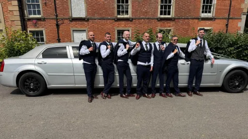 Group of groomsmen standing beside a silver Chrysler stretch limousine parked in front of a red-brick wedding venue in Antrim.