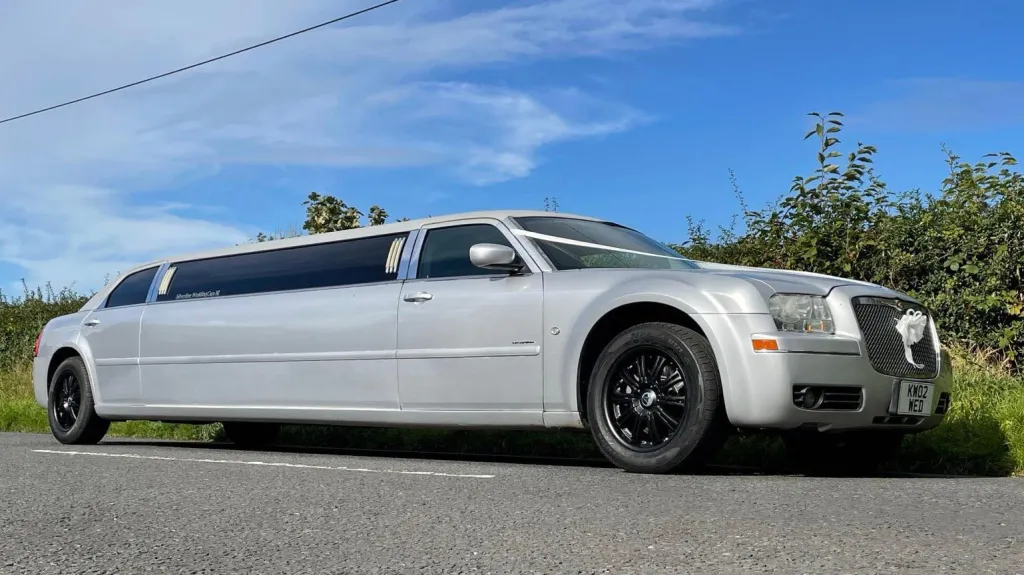 Side view of a silver Chrysler stretch limousine on a country road in Northern Ireland, showing its extended body, black alloy wheels and chrome grille, dressed with ribbons and bows on front bonnet.