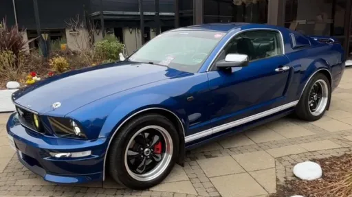 Left side profile of a blue Ford Mustang with sporty lines and chrome wheels, parked outside a modern venue.