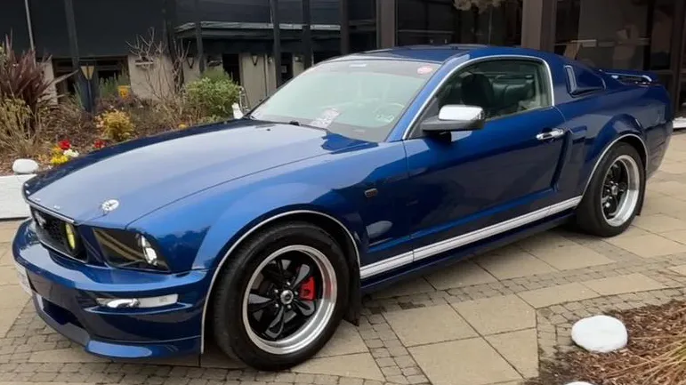 Left side profile of a blue Ford Mustang with sporty lines and chrome wheels, parked outside a modern venue.