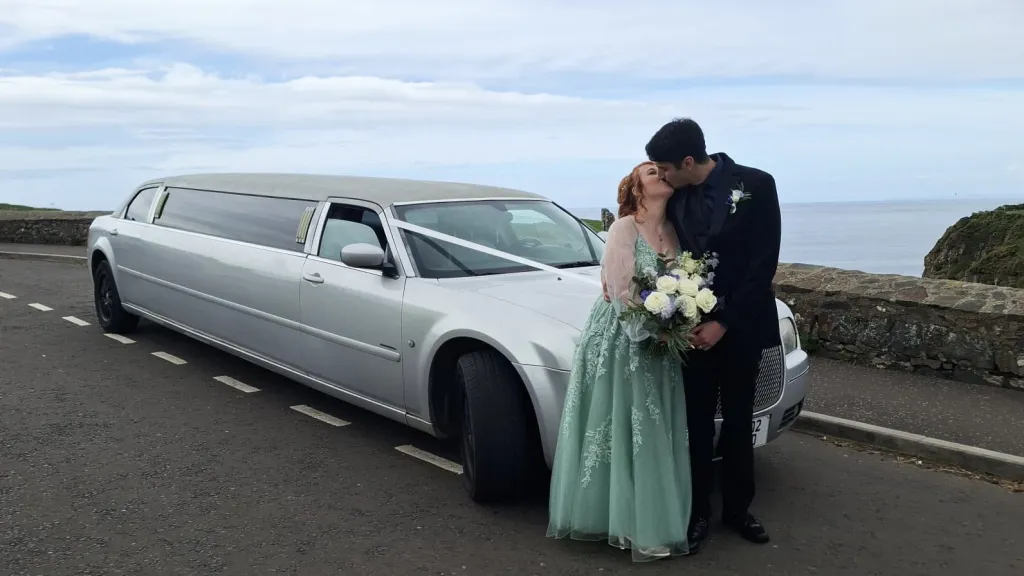 Bride and groom beside a silver Chrysler stretch limousine parked near the coast in Belfast.