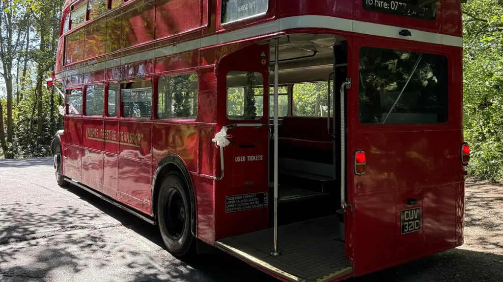 Rear view of a red Routemaster double-decker bus with open platform and vintage design, parked on a tree-lined street.