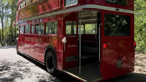 Rear view of a red Routemaster double-decker bus with open platform and vintage design, parked on a tree-lined street.
