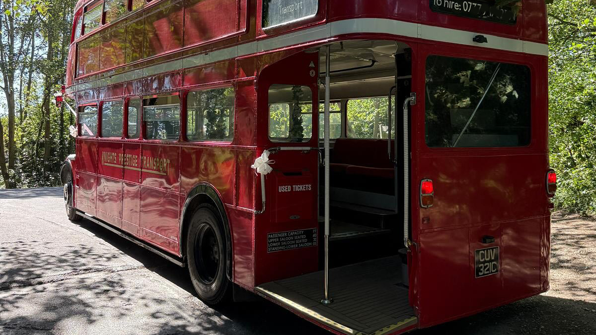 Rear view of a red Routemaster double-decker bus with open platform and vintage design, parked on a tree-lined street.