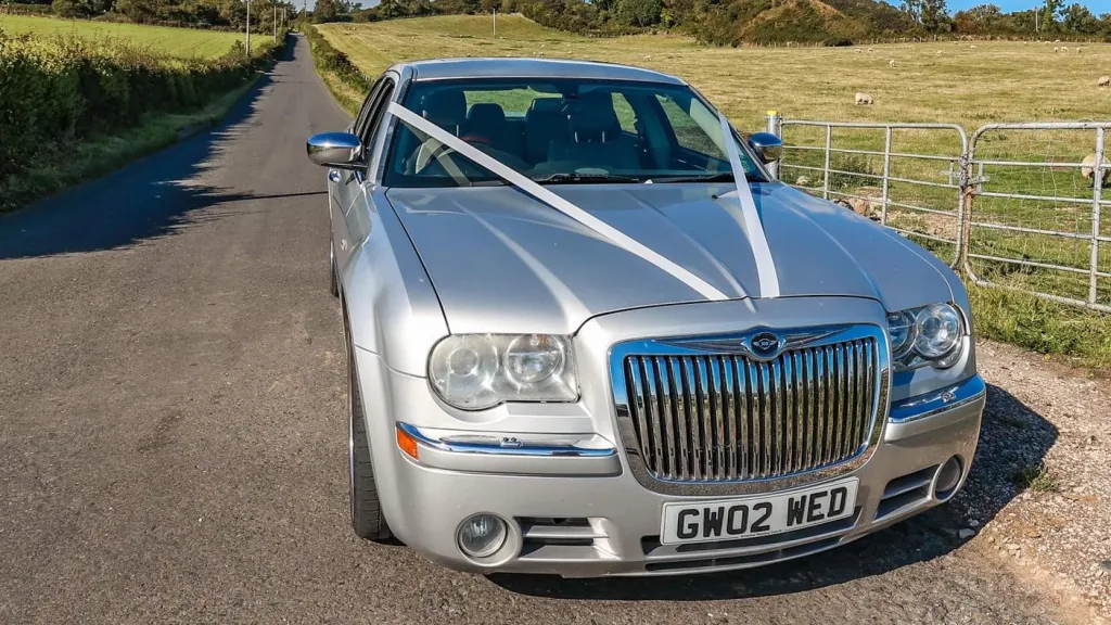 Angled front view of a silver Chrysler 300C saloon decorated with white wedding ribbons, parked beside open countryside.