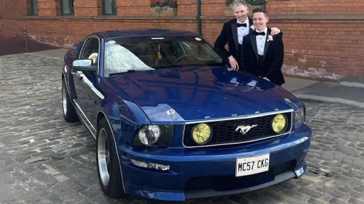 Two grooms posing with a blue Ford Mustang wedding car parked outside a red-brick venue in Northern Ireland.