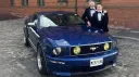 Two grooms posing with a blue Ford Mustang wedding car parked outside a red-brick venue in Northern Ireland.