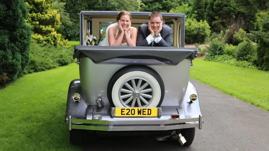 Bride and groom seated inside a silver Imperial convertible wedding car with the roof lowered, smiling while driving through the countryside.