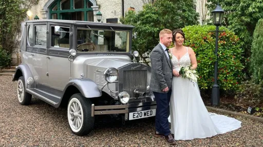 Bride and groom posing beside a silver Imperial vintage wedding car with white ribbons, parked in front of a grand venue.