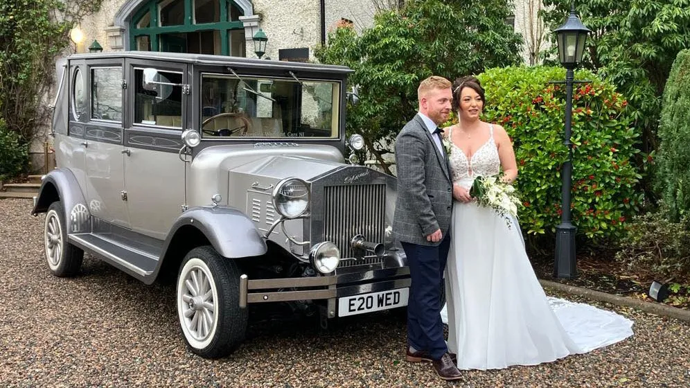 Bride and groom posing beside a silver Imperial vintage wedding car with white ribbons, parked in front of a grand venue.