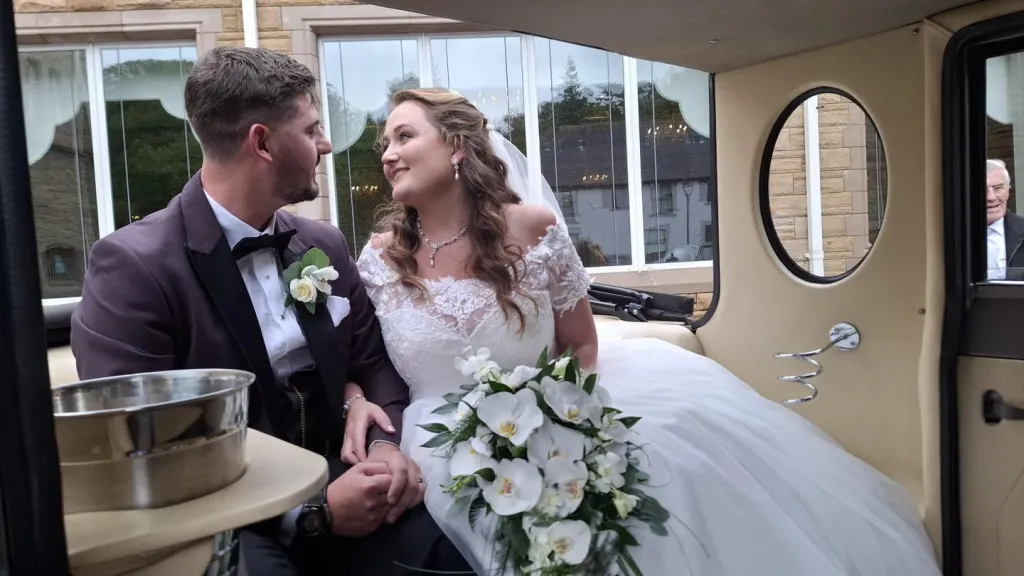 Bride and groom seated inside a cream leather interior of an Imperial wedding car, with sunlight shining through the rear windows.