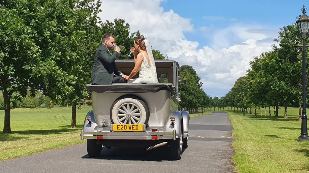 Bride and groom enjoying champagne inside a silver Imperial convertible wedding car while parked in a scenic parkland setting.