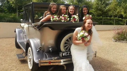 Bride and bridesmaids posing beside a silver Imperial convertible wedding car with white ribbons and floral decorations.