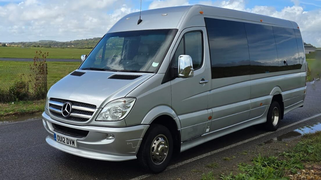 Front left view of a silver Mercedes 16-seater minibus with large tinted privacy windows, parked on a countryside road in Sussex.