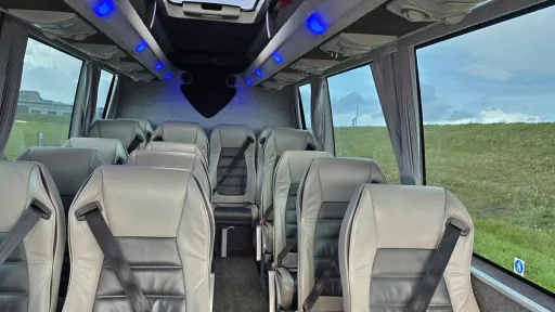 Interior of a Mercedes 16-seater minibus featuring silver-grey leather seats, overhead storage, and wide windows for passenger comfort.