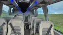 Interior of a Mercedes 16-seater minibus featuring silver-grey leather seats, overhead storage, and wide windows for passenger comfort.