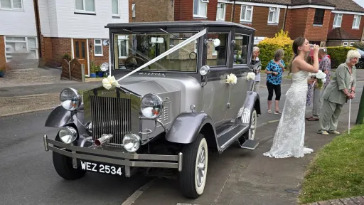 Silver Imperial wedding car decorated with white ribbons, parked in front of a wedding venue with guests in the background.