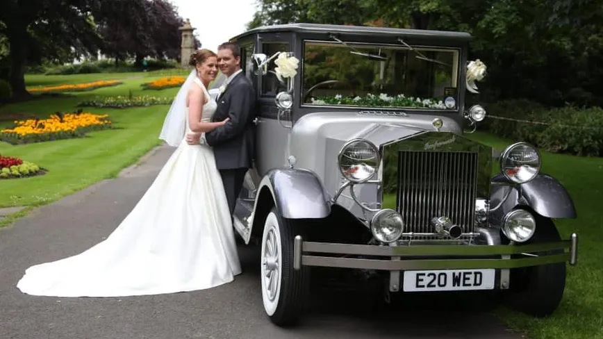 Bride and groom embracing beside a silver Imperial wedding car decorated with ribbons, parked on a country driveway.