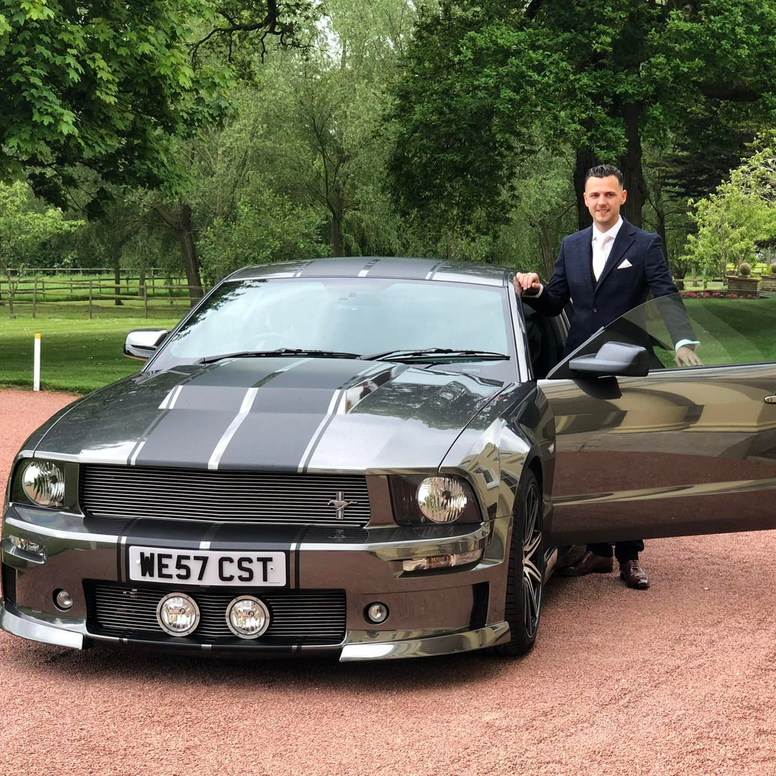 Groom standing beside a silver American Ford Mustang wedding car with black racing stripes, parked on a red gravel driveway surrounded by greenery.