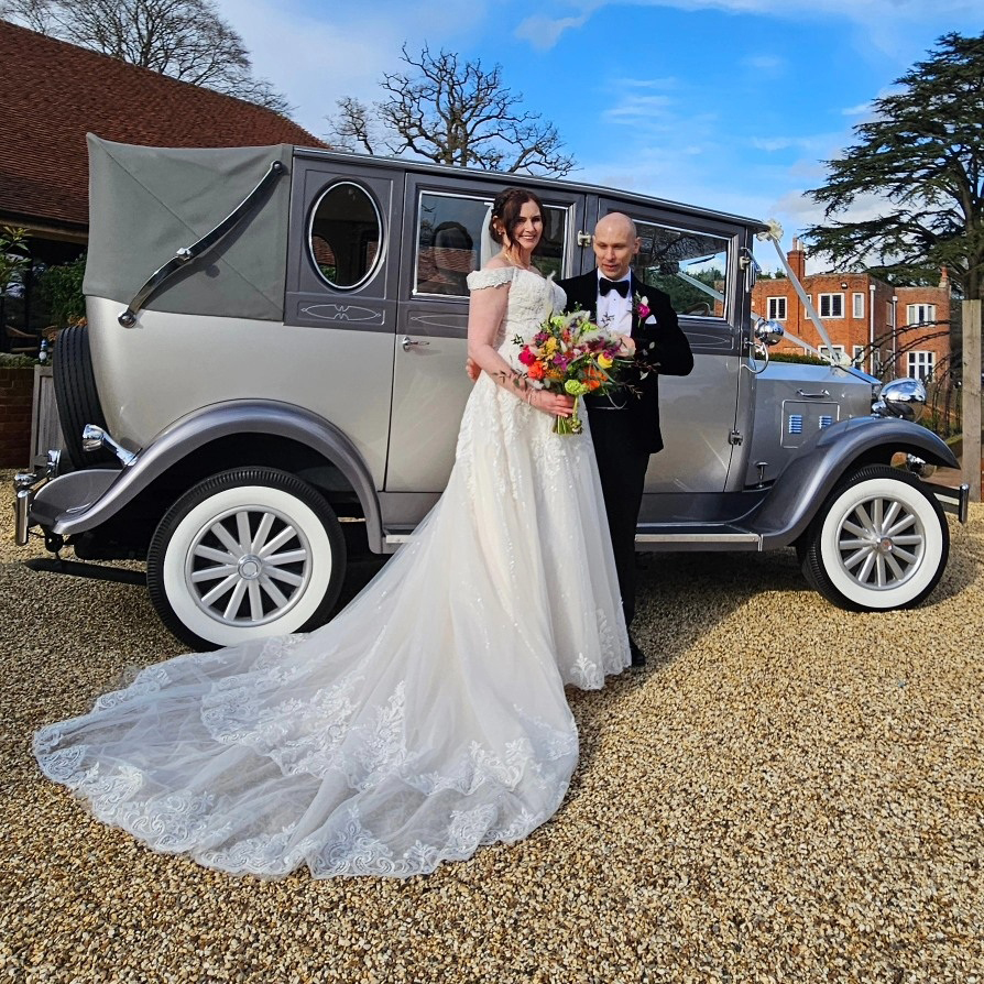 Bride and groom posing in front of a silver vintage Imperial-style wedding car outside a countryside venue on a sunny day.