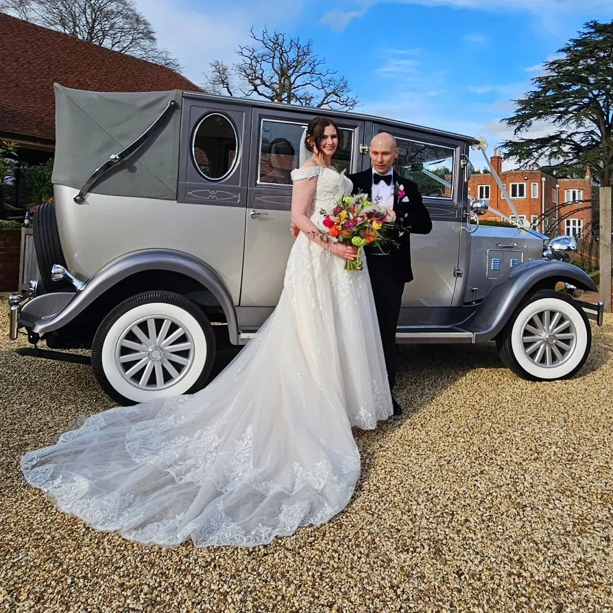 Bride and groom posing in front of a silver vintage Imperial-style wedding car outside a countryside venue on a sunny day.