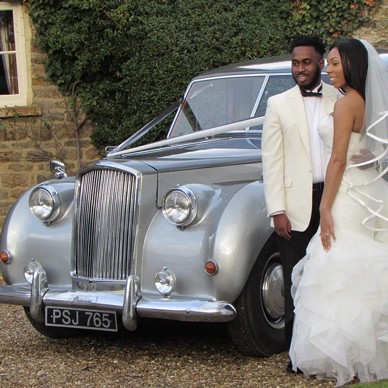Bride and groom standing together beside a silver classic limousine with vintage curves outside a traditional stone building.