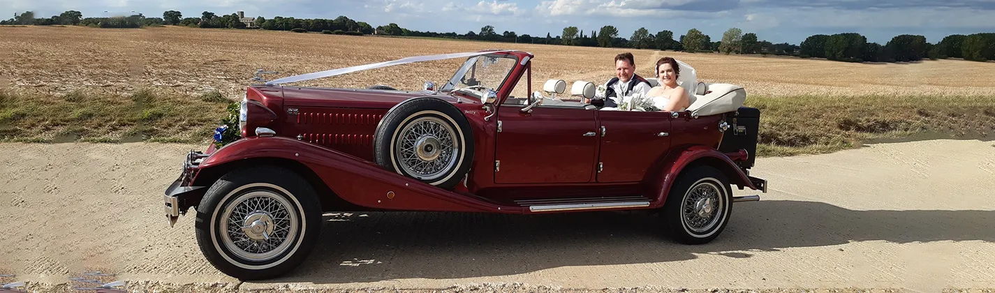 Left sode view of a Burgundy Red vintage-style Beauford with convertible roof open and couple seating in the rear seat.
