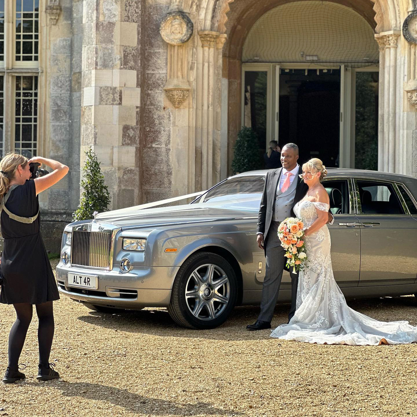 Newlyweds with a modern silver Rolls-Royce Phantom wedding car parked at a grand manor entrance, captured by their photographer.