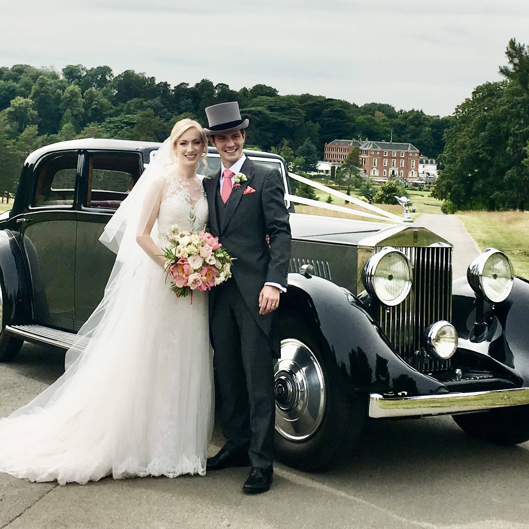Bride and groom standing beside a grey vintage Rolls-Royce wedding car with classic lines, set against a green park backdrop.