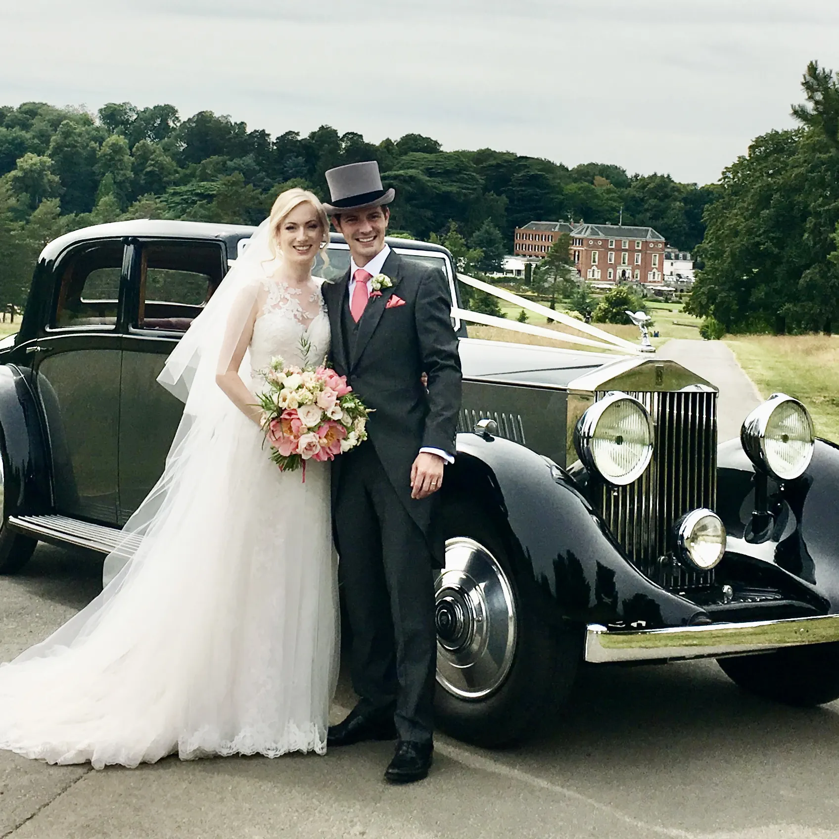 Bride and groom standing beside a grey vintage Rolls-Royce wedding car with classic lines, set against a green park backdrop.
