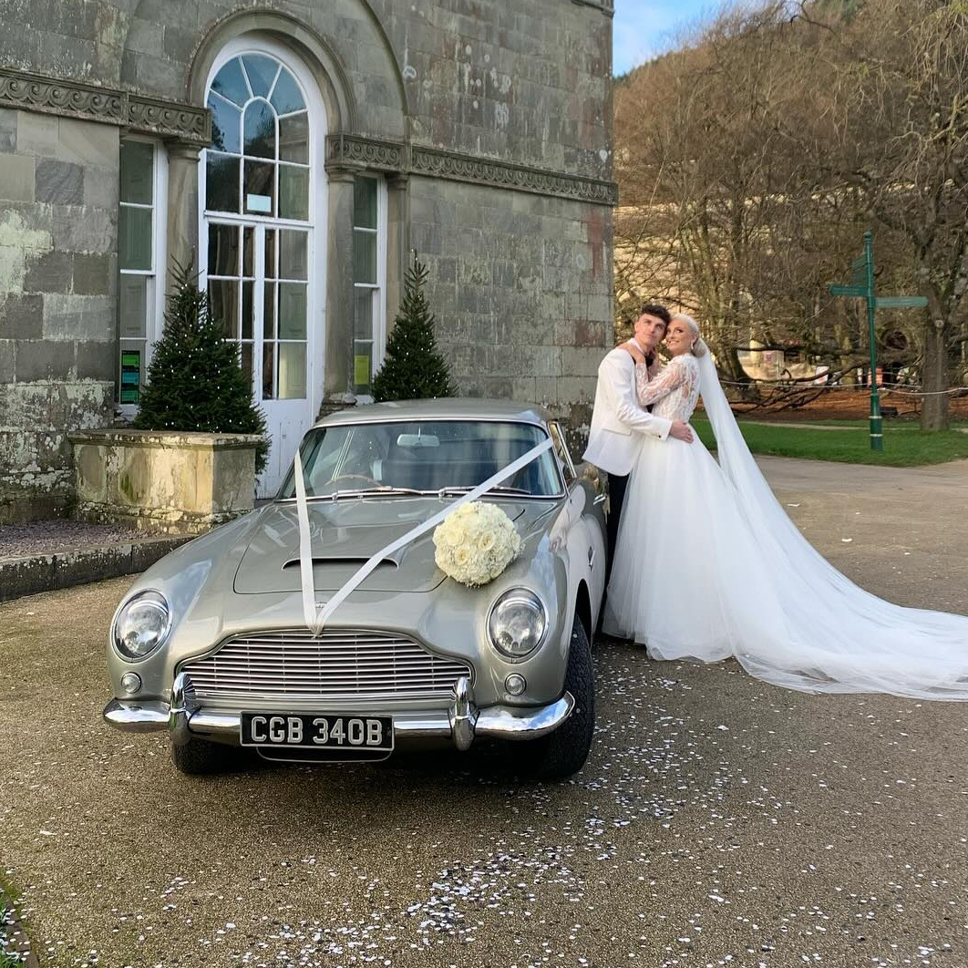 Bride and groom beside a silver Aston Martin DB5 wedding car with white ribbons, outside a stately home.
