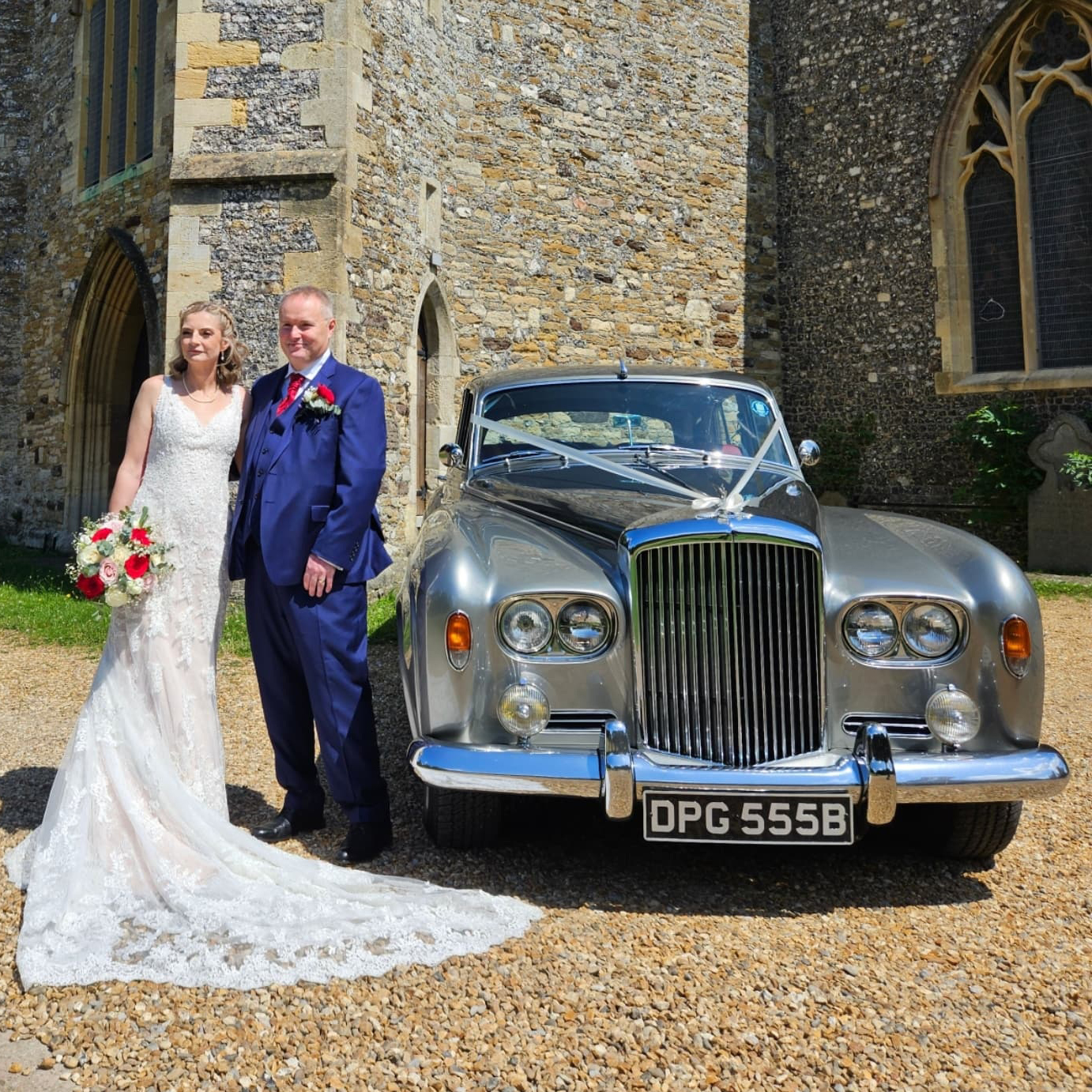 Smiling couple standing next to a silver classic Bentley wedding car outside a rustic church entrance.