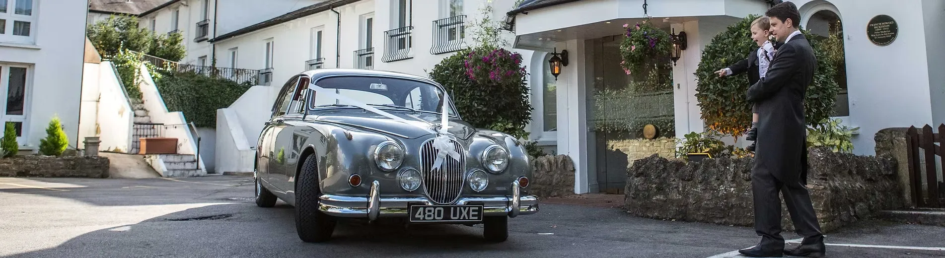 A Silver Grey Classic Jaguar Mk2 dressed with white ribbons parked in front of a wedding venue with the groom standing in front of the vehicle holding his son who is pointing at the silver car.