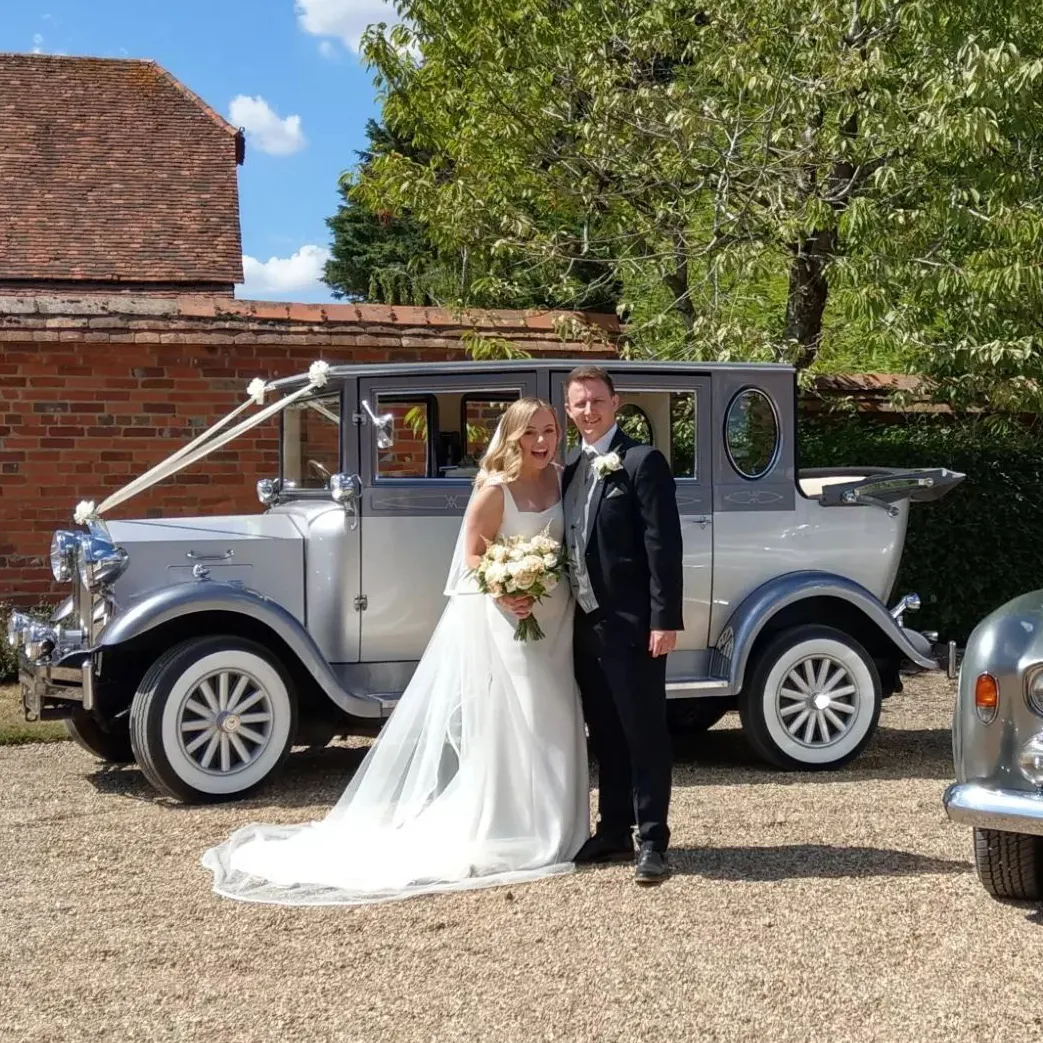 Elegant couple posing beside a silver vintage wedding car with whitewall tyres and classic styling in a sunny courtyard.