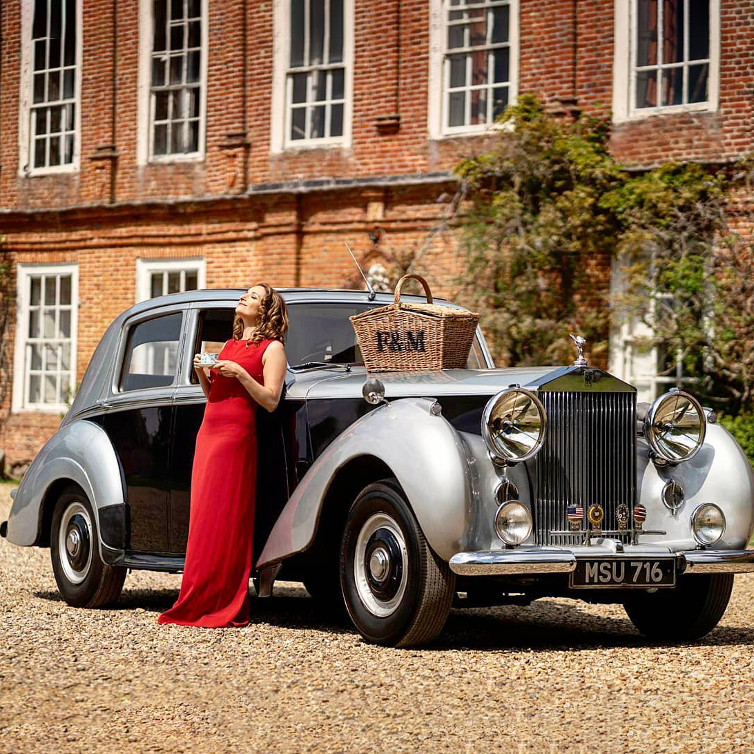 Bridesmaid in a red dress with a vintage silver Rolls-Royce wedding car outside a Georgian-style venue.
