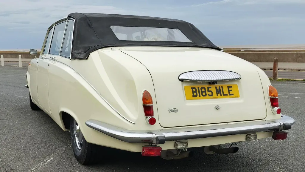 Angled rear left view of a classic ivory Daimler DS420 limousine with chrome bumpers and soft-top convertible roof closed.