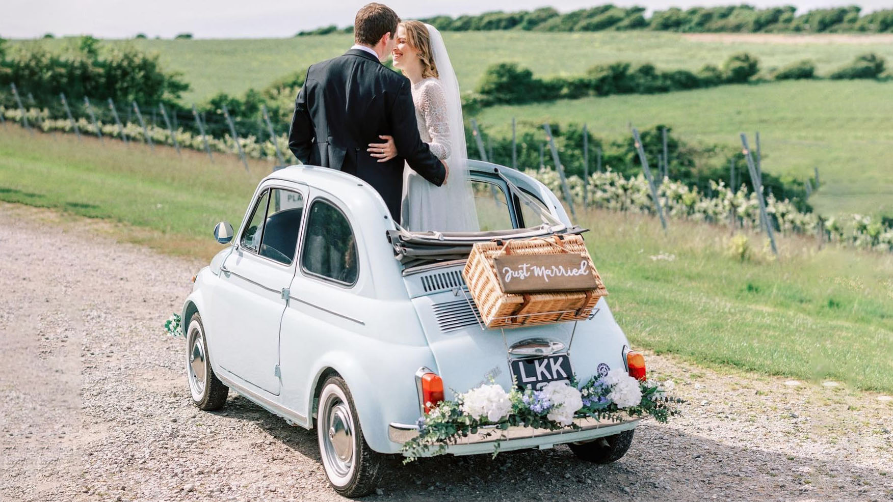 ride and groom sharing a romantic moment in a pale blue Fiat 500 convertible with “Just Married” sign on the back and floral wedding decoration attached to the rear chrome bumper