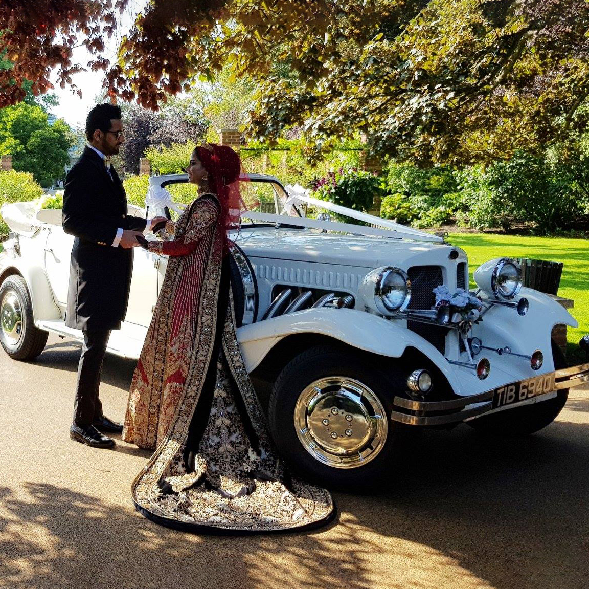 Asian bride and groom standing in front a white vintage Beauford convertible in a park. Bride is wearing a traditional red and black Asian ceremonial dress.