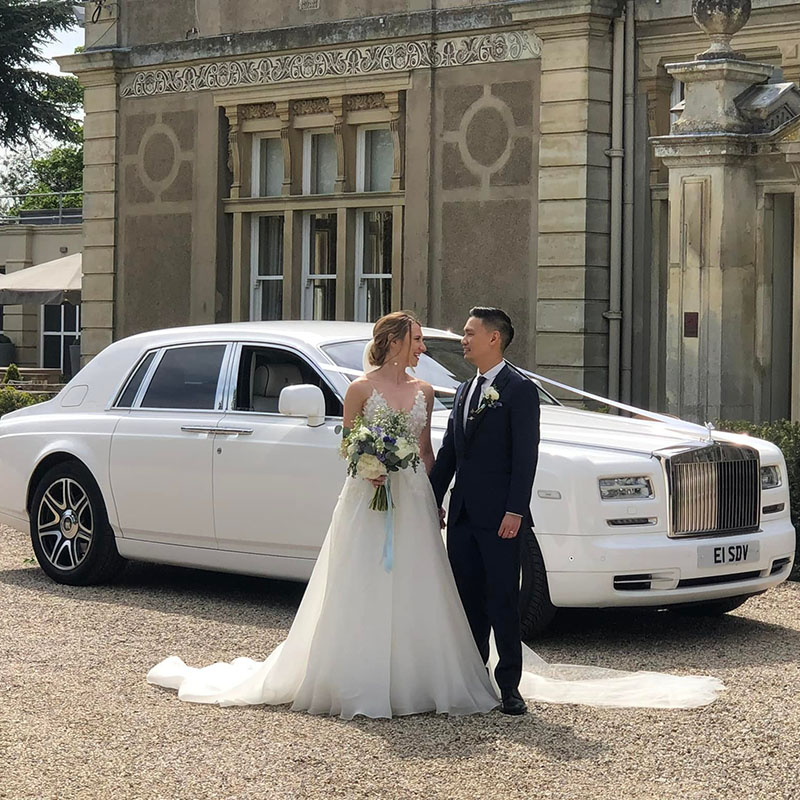 Bride wearing a white wedding gown and holding a colourful bouquet of flower standing on the right of her groom in front of a luxurious white Rolls-Royce Phantom parked outside a grand wedding venue.