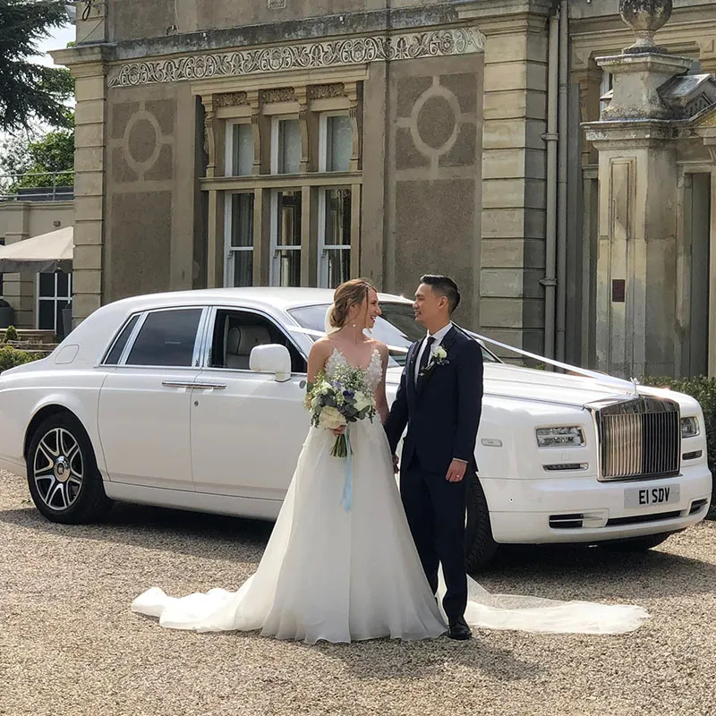 Bride wearing a white wedding gown and holding a colourful bouquet of flower standing on the right of her groom in front of a luxurious white Rolls-Royce Phantom parked outside a grand wedding venue.