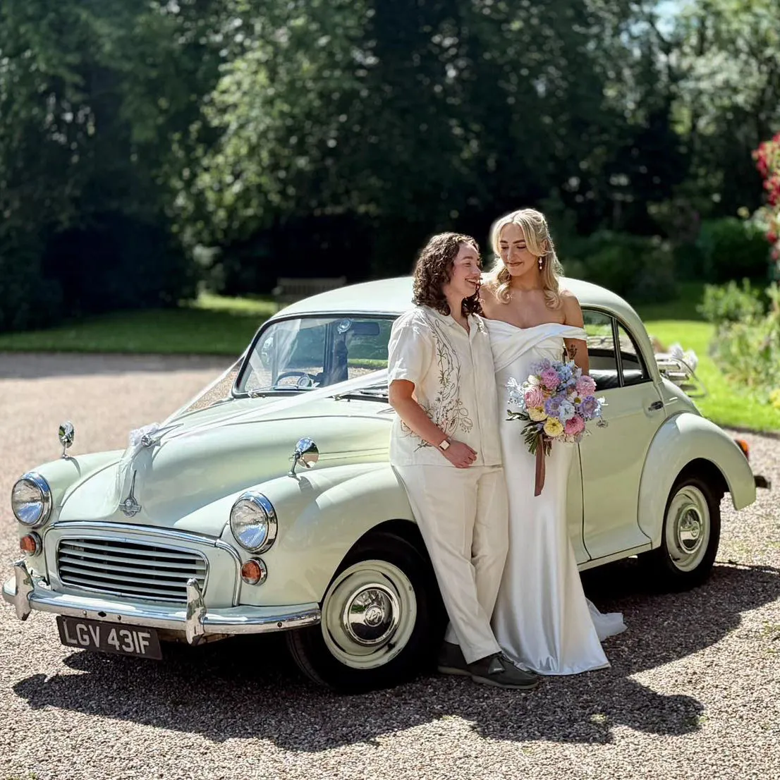 Two smiling brides, posing in front of an Ivory classic Morris Minor wedding car at an LGBTQIA+ wedding. Bride on the right of the photo is holding a colourful wedding bouquet of blue, pink and yellow flowers.