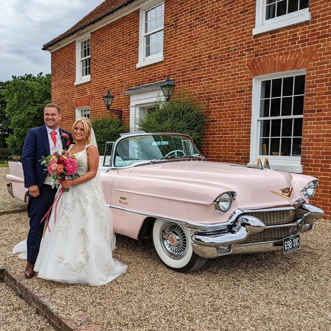 Bride and groom smiling beside a vintage pink American convertible wedding car. The car has white wall tires and large chrome bumpers. Groom is wearing a blue suit with an orange tie and Bride is wearing a white designer wedding dress.
