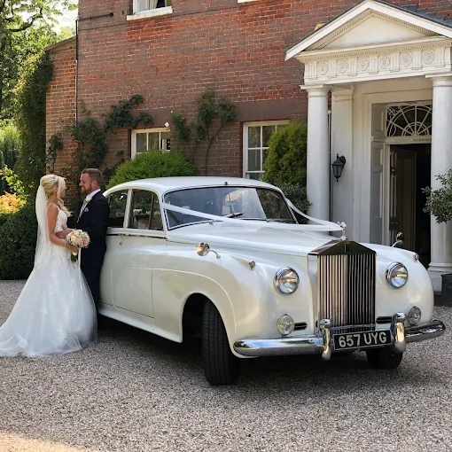 Asian bride and groom standing in front a white vintage Beauford convertible in a park. Bride is wearing a traditional red and black Asian ceremonial dress.