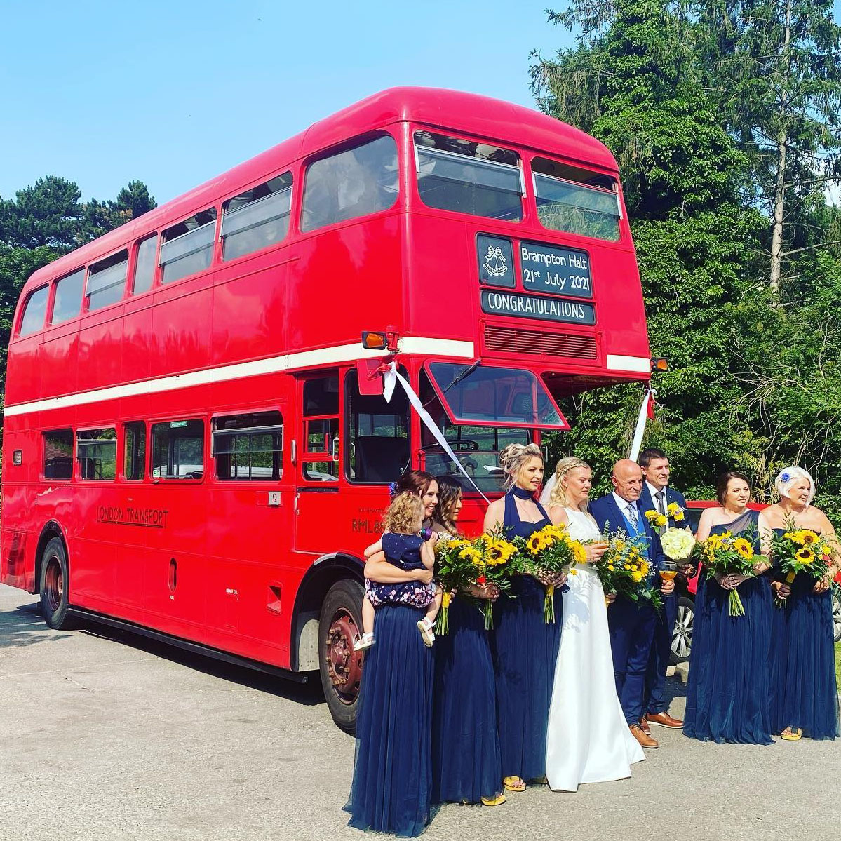 Wedding party posing with a classic red Routemaster double-decker bus at wedding venue.  Wedding party are wearing navy blue dresses and suits and holding with yellow matching sunflowers bouquet and the bride is standing in the middle in her white dress.