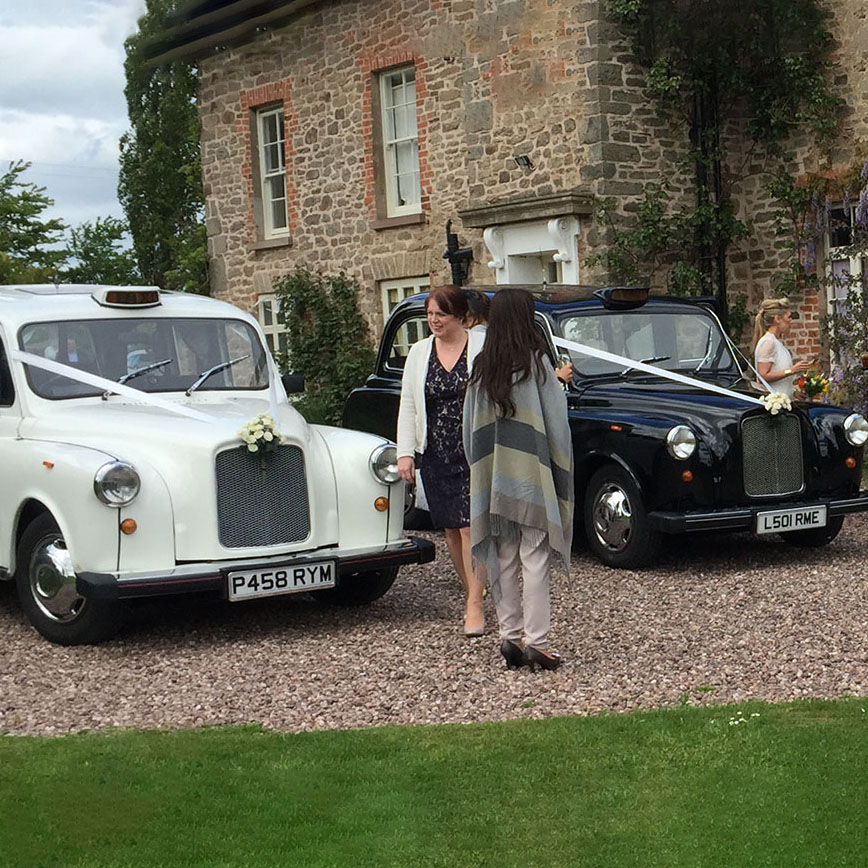 Classic Taxi cabs on wedding duties. Left taxicab is white and right one is black. Both dressed in matching white ribbons across front bonnet with added ivory bouquet of flowers on top of their front grill.