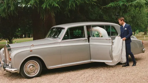 Bride stepping into a classic silver Rolls-Royce wedding limousine outside a country venue.