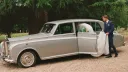 Bride stepping into a classic silver Rolls-Royce wedding limousine outside a country venue.