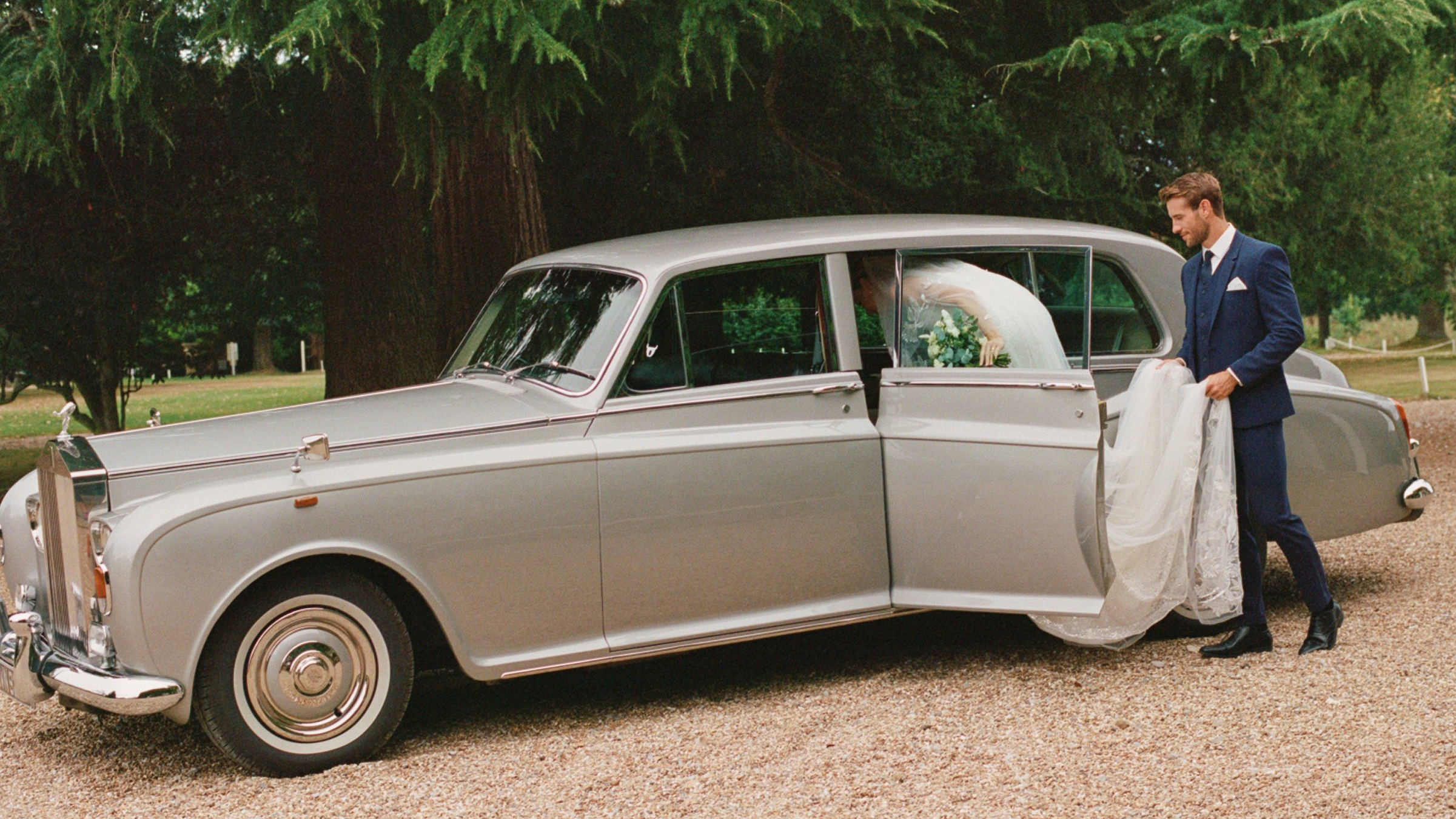 Bride stepping into a classic silver Rolls-Royce wedding limousine outside a country venue.